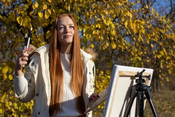Fototapeta na wymiar beautiful, red-haired girl with long hair draws on an easel in the park, on a sunny day