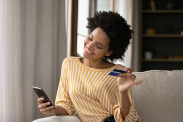 Happy young African consumer woman excited with shopping on internet, using ecommerce app on mobile phone, paying for purchase, order by credit card, buying goods online, resting on sofa at home