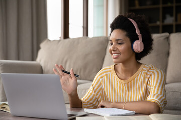 Happy African American student girl in wireless headphones studying online at computer at home,...