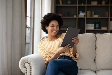 Happy young Black girl relaxing on sofa, using tablet computer, reading book, watching movie, media...