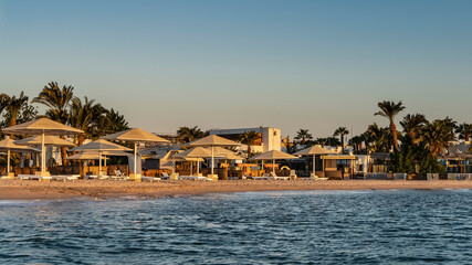 Morning on the Red Sea beach. There are empty sunbeds and chairs on the sand under the sun umbrellas. Ripples on the blue water. Palm trees against the azure sky. Egypt