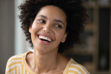 Happy joyful Black girl looking away with toothy smile, laughing. Beautiful young curly haired woman, model, dental clinic patient with perfect white teeth posing indoors. Candid home portrait