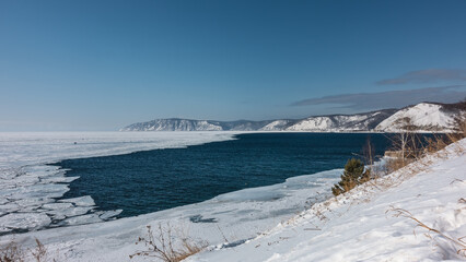 The ice-free Angara River flows out of the frozen Lake Baikal. Melted ice floes on blue water. Tiny silhouettes of people on the ice. Snow and dry grass on the shore. Mountains against the azure sky.