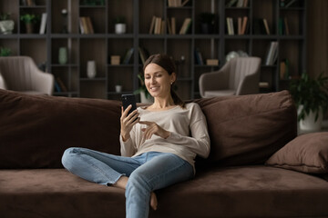 Fototapeta premium New mobile application ad, videoconferencing call event, weekend activity free time with modern tech usage concept. Young woman spend leisure at home holds smart phone while relax alone in living room