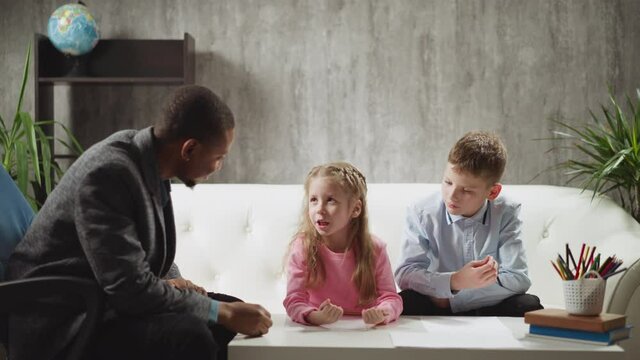 Cute little girl student tells new words to African-American teacher while brother sits nearby on sofa at home lesson with native speaker slow motion