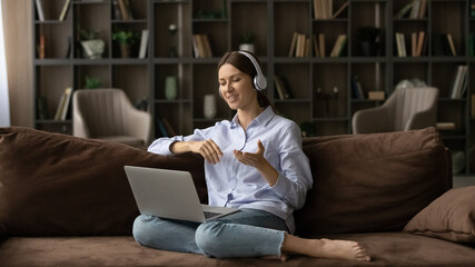 Young woman sit on couch in living room put laptop on laps wear modern headphones talk to family living abroad using video call application. Virtual meeting, easy comfort remote communication concept