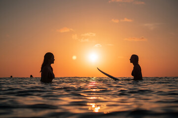 Portrait from the water of two surfer girls with beautiful bodies on surfboards in the ocean at colourful sunset time