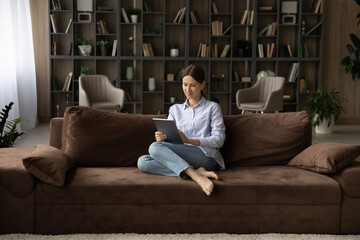 Young woman relax on comfy sofa with tablet buying goods and items for home on internet. Client of...
