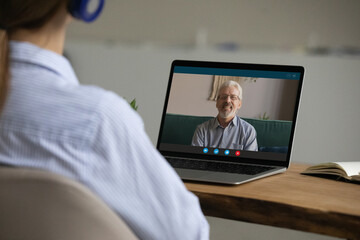 Fototapeta premium Rear view woman sit at table wear headphones talk to elderly happy father via video conference app, laptop screen view over female shoulder. Virtual meeting, modern tech, connection, videocall concept