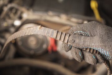 An auto mechanic holds in his hand a torn timing belt with worn out teeth against the background of a car engine - obrazy, fototapety, plakaty