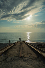 A man stands on a concrete pier going into the sea. On the water track from the evening sun. Blue sky with beautiful clouds. A woman goes swimming in the sea.