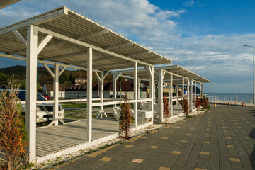 A seafront with white wooden awnings and paving tiles. In the background is a blue sky with clouds....