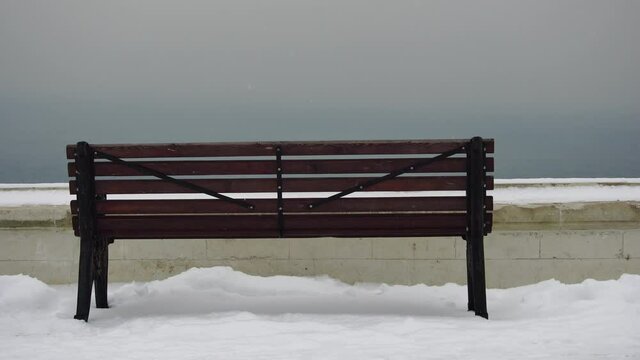 Snow is falling on empty city park. A lonely snow-covered bench stands near sea. Christmas Winter New Year Scenery background. Frosty winter in the city after snowfall