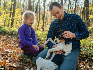 A man in plaid coffee and jeans is stroking a dog lying on his lap. A girl sits next to him and looks at them. Father and daughter in the autumn forest. The concept of a happy family.