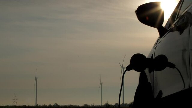 Wind turbines produce alterative energy in field. Sunlight shines over electric car charging at rural station against rotating propellers close view