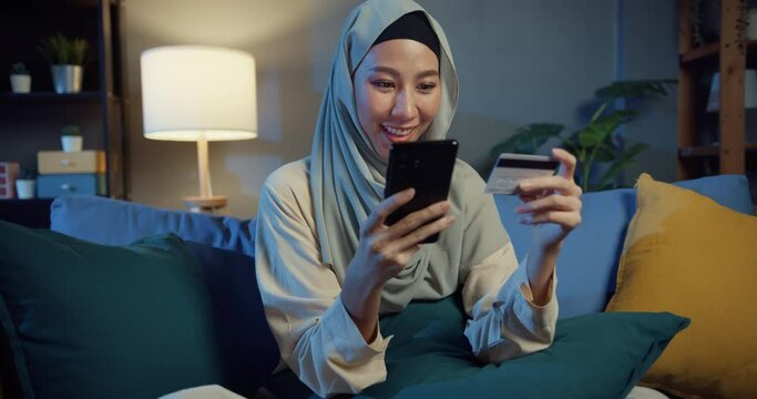 Attractive cheerful young Asia islam muslim beauty girl in hijab with casual smile sitting on couch use phone enjoy successful electronic online shopping with credit card in cozy living room at night.