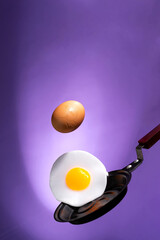 whole and fried eggs above a frying pan float in the air on a violet. Breakfast time