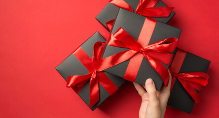 First person top view photo of hand holding black giftbox with red ribbon bow over stack of gift...