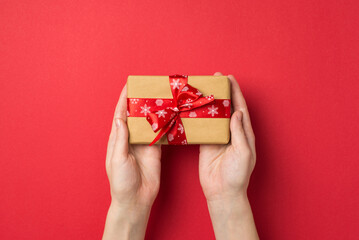First person top view photo of female hands holding craft paper giftbox with red ribbon bow on isolated red background