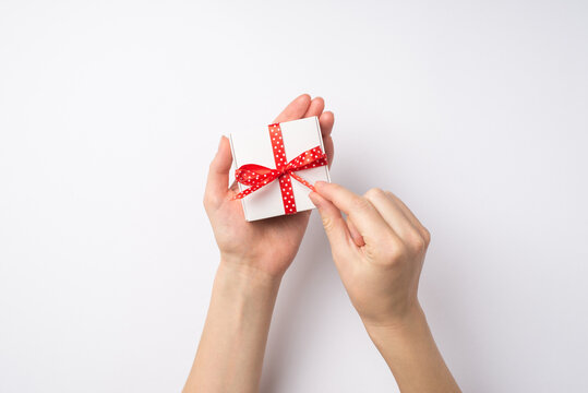 First person top view photo of young woman's hands unpacking small white giftbox with red dotted ribbon bow on isolated white background