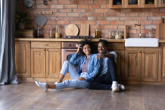 Relaxed joyful young African American family couple sitting on warm heated wooden floor in stylish cottage house kitchen, spending weekend time in own dwelling, real estate concept. - Powered by Adobe