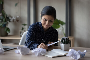 Stressed anxious young Indian businesswoman feeling nervous, creating new ideas or doing paperwork, sitting at table with crumpled documents or sheets with notes at workplace, deadline concept.
