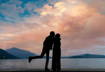 Fototapeta premium Silhouette of a cute couple in love at the moment of kissing with a background of a lake at sunset with clouds of warm tones illuminated by the sun.