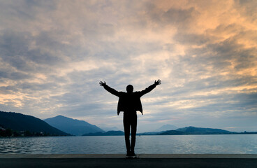 Silhouette of a man with his back to camera, arms outstretched in front of a landscape of a lake at...