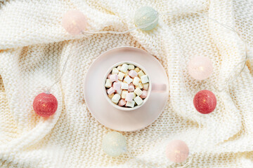 Pink cup of cocoa with marshmallows on white knitted plaid with light garlands. Winter holidays concept. Top view, flat lay
