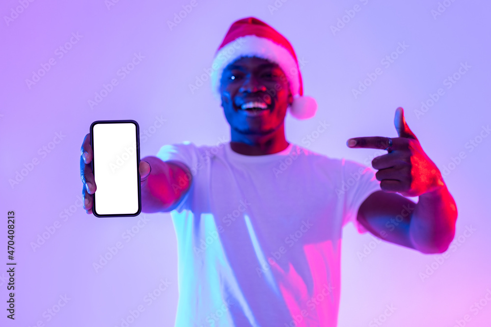 Poster smiling young black man in santa hat pointing at empty smartphone screen in neon light, mockup for m
