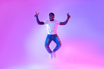 Full length portrait on young African American man jumping and showing peace gesture in neon light