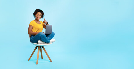 Positive young black woman sitting on chair with laptop, showing thumb up gesture, recommending online work or studies