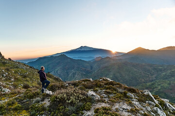 Man standing on top of the cliff in the mountains at sunset enjoying the beautiful sunset over erupting volcano Etna. Beautiful view in Sicily, Italy