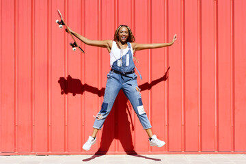 Black woman dressed casual, wtih a skateboard jumping for joyon red urban wall background.