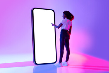 Young black woman touching screen of giant smartphone in neon light, mockup for new mobile app or website - obrazy, fototapety, plakaty