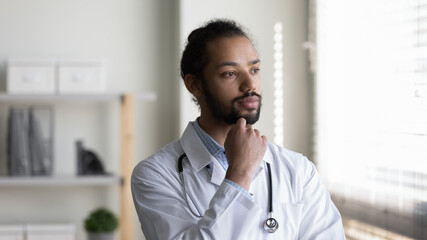 Head shot of pensive young African American man doctor looking in distance, touching chin, thoughtful serious physician general practitioner surgeon in white uniform thinking and visualizing