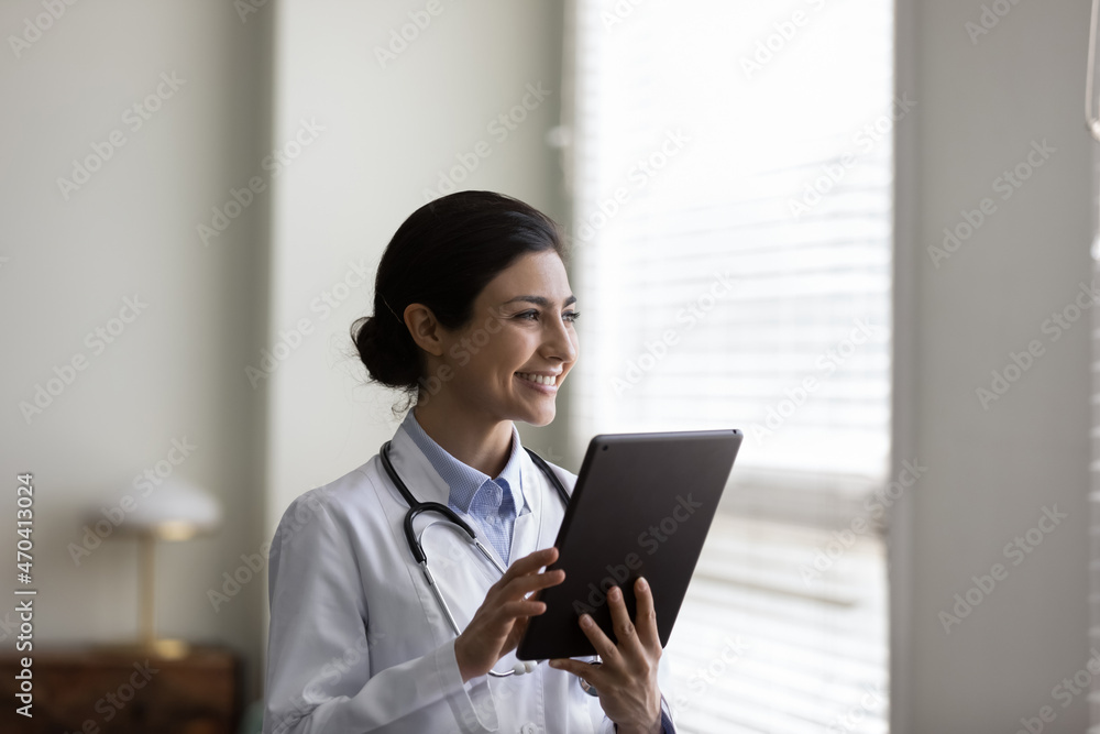 Wall mural smiling dreamy young indian woman doctor physician in white uniform with stethoscope holding tablet,