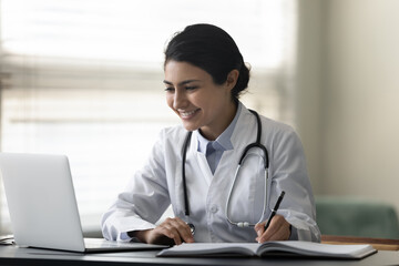 Smiling Indian female doctor physician in uniform with stethoscope using laptop, taking notes,...