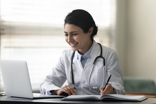 Smiling Indian female doctor physician in uniform with stethoscope using laptop, taking notes, writing in medical journal, professional therapist practitioner filling documents or watching webinar