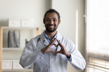 Head shot portrait smiling young African American man doctor in white uniform with stethoscope showing heart gesture, looking at camera, standing in hospital office, expressing support and care