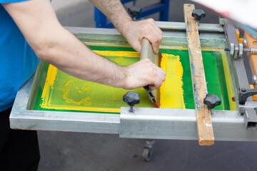 Serigraphy silk screen print process at clothes factory. Frame, squeegee and plastisol color yellow paints
