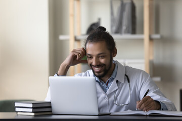 Smiling African American man doctor intern using laptop, looking at screen watching webinar, taking...