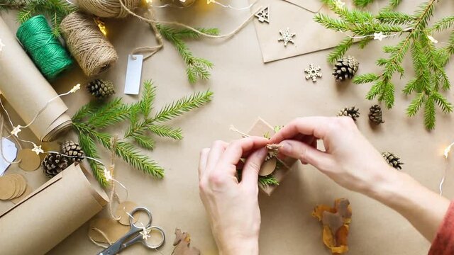 Hands pack a gift for Christmas and new year in eco-friendly materials: kraft paper, live fir branches, cones, twine, tied with a bow. Tags with mock up, natural decor, hand made, flatly. Festive mood