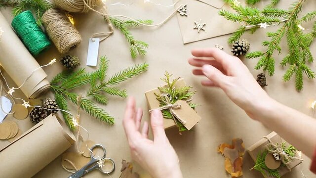 Hands pack a gift for Christmas and new year in eco-friendly materials: kraft paper, live fir branches, cones, twine, tied with a bow. Tags with mock up, natural decor, hand made, flatly. Festive mood