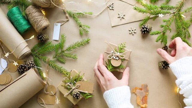 Hands pack a gift for Christmas and new year in eco-friendly materials: kraft paper, live fir branches, cones, twine, tied with a bow. Tags with mock up, natural decor, hand made, flatly. Festive mood