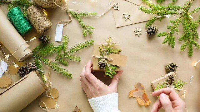 Hands pack a gift for Christmas and new year in eco-friendly materials: kraft paper, live fir branches, cones, twine, tied with a bow. Tags with mock up, natural decor, hand made, flatly. Festive mood