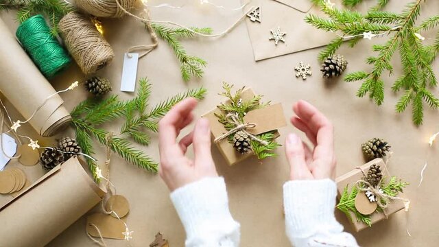 Hands pack a gift for Christmas and new year in eco-friendly materials: kraft paper, live fir branches, cones, twine, tied with a bow. Tags with mock up, natural decor, hand made, flatly. Festive mood