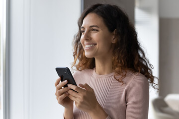 Head shot smiling dreamy woman distracted from modern smartphone, looking in distance, chatting online with boyfriend, waiting for message or phone call, enjoying leisure time with mobile device