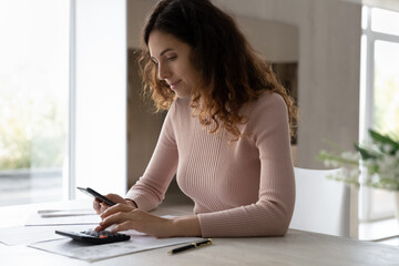 Young woman calculating expenses, paying taxes, using smartphone and calculator, sitting at desk...