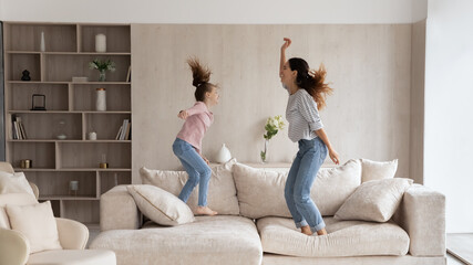 Overjoyed mother with little daughter having fun, jumping on couch at home, happy young mom with...